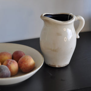 An antique ceramic pitcher with a glossy glaze in an cream and beige color palette, displayed next to a bowl of fruits on a dark table.