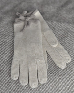 A pair of beige cashmere gloves with a bow detail on the wrist, displayed on a grey background.