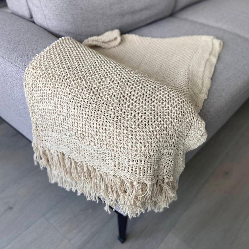 A beige knitted cotton throw with tassels draped over the edge of a gray sofa, showcasing its soft texture and elegant design.