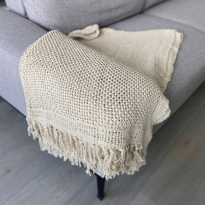 A beige knitted cotton throw with tassels draped over the edge of a gray sofa, showcasing its soft texture and elegant design.