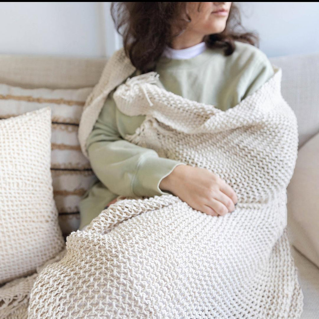 Person wrapped in a beige knitted blanket sitting on a couch.