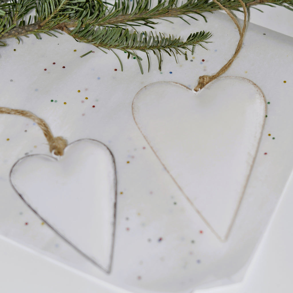 Two white metal heart-shaped ornaments with jute hanging loops, one larger than the other, displayed against a white background.