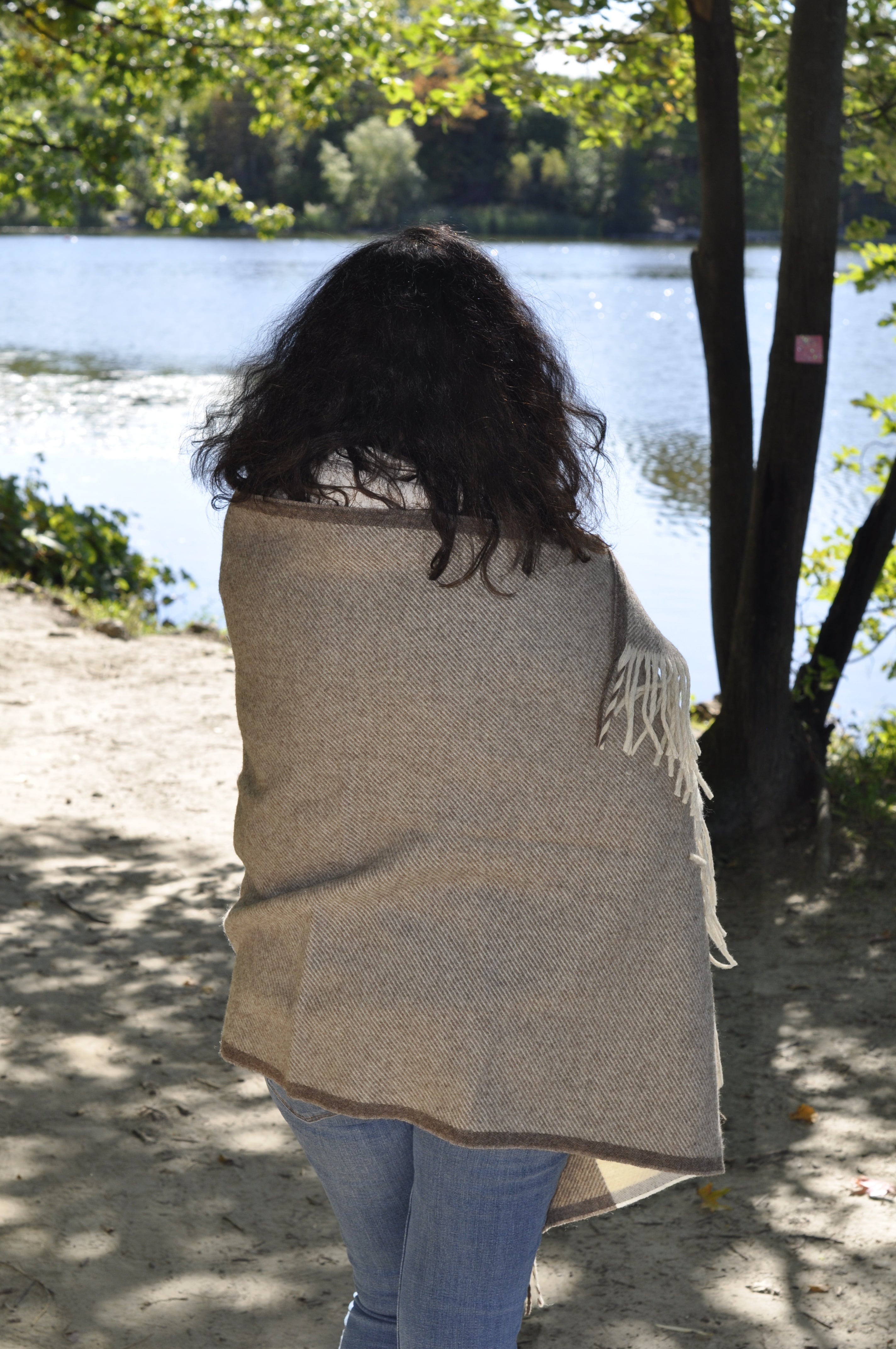 Person wearing a beige shawl standing outdoors with trees and water in the background