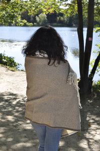 Person wearing a beige shawl standing outdoors with trees and water in the background
