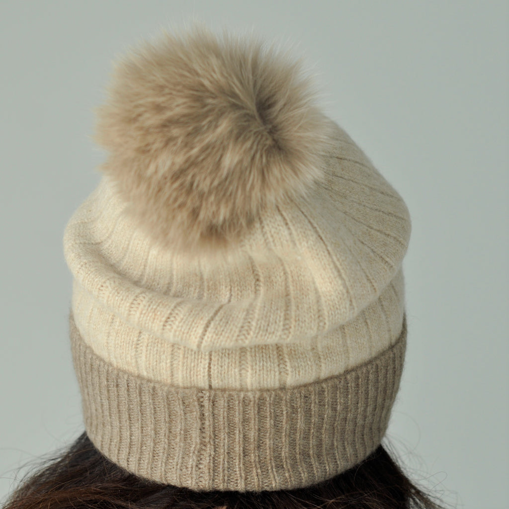 A beige cashmere pom pom beanie hat with a fluffy pom pom on top, made of a ribbed design, displayed on a mannequin.