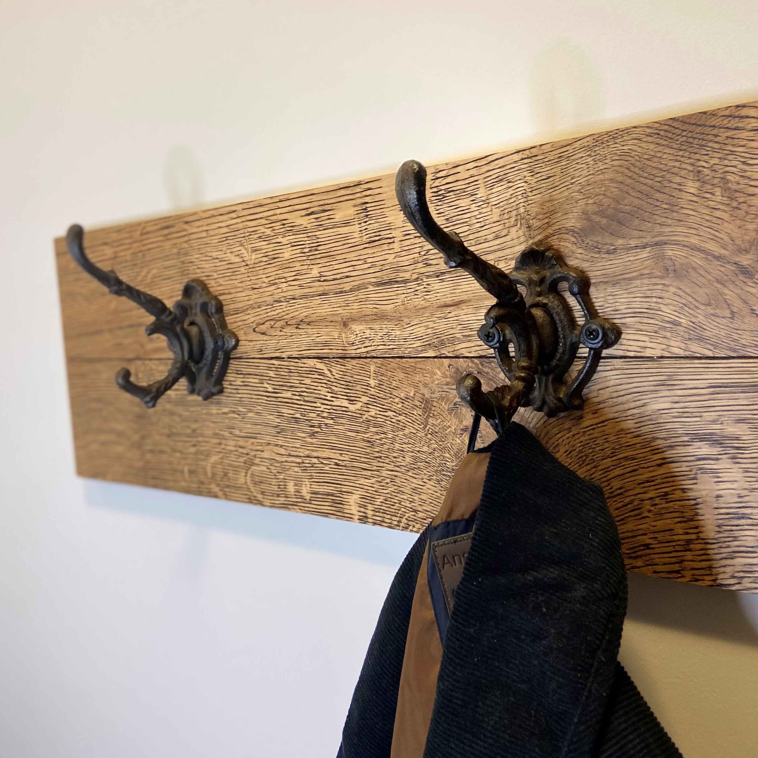 A cast iron double hook coat rack mounted on a wooden panel, with dark brown iron hooks and a brown wooden background, holding a dark jacket and a hat.