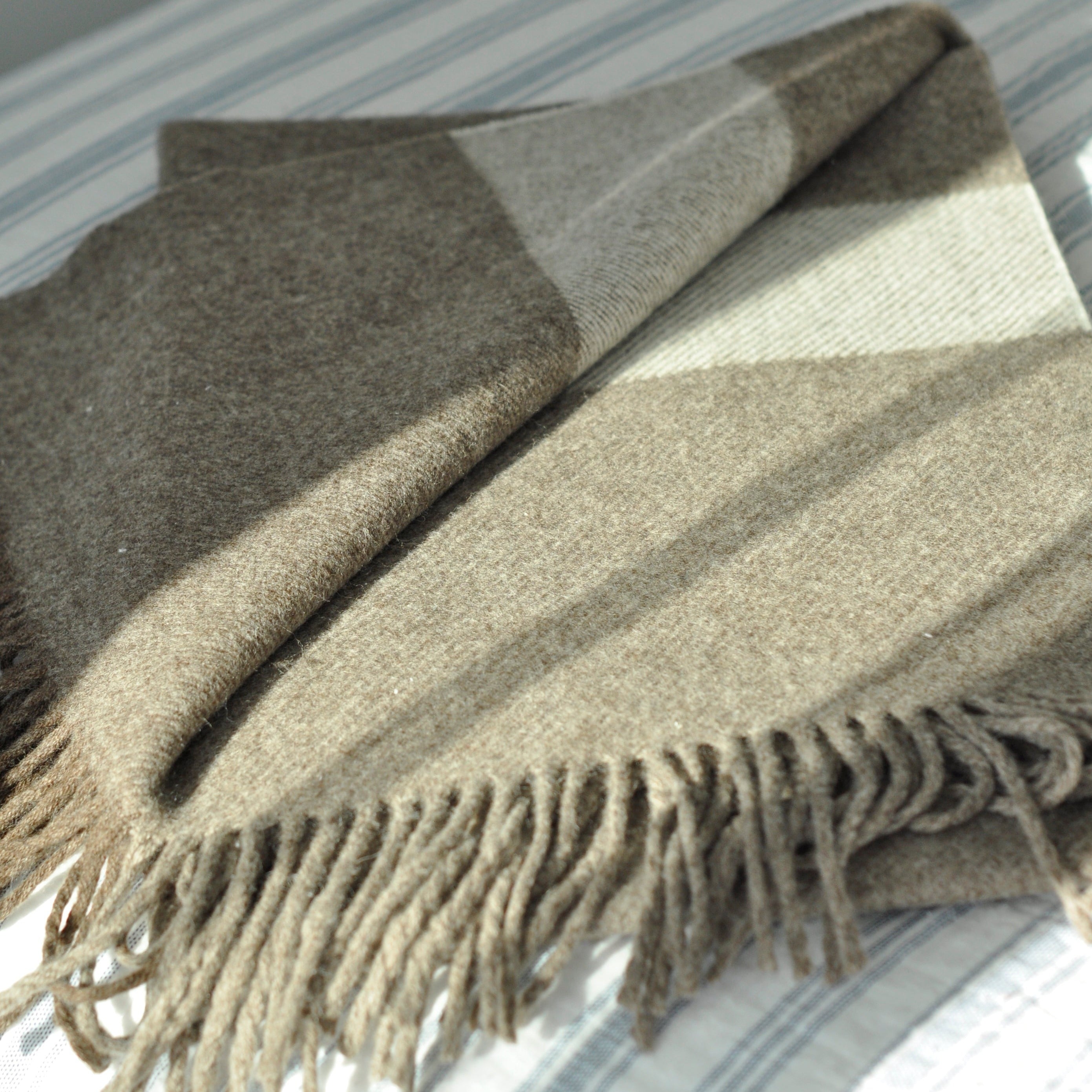 A chocolate brown wool scarf with long tassels, laid out on a bed with a striped background.